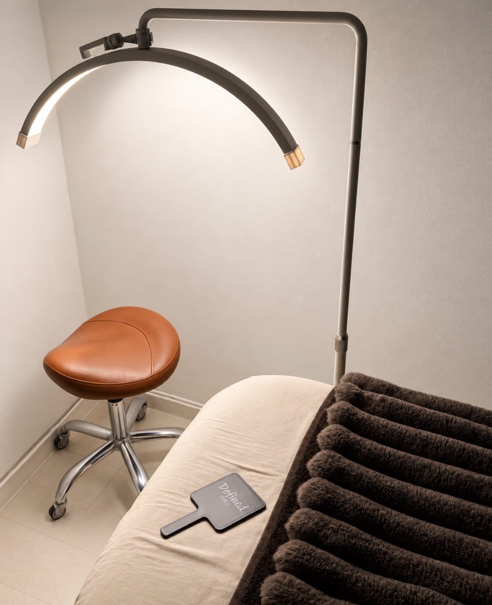 The Defined PMU treatment room in Punavuori: a reclined bed under an arc lamp, a tan leather stool, and a Defined hand mirror on the bed